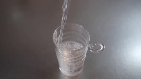Above-angled-full-shot-of-water-being-poured-into-a-glass-cup-on-a-metallic-kitchen-worktop,-health-and-clean-water-concept,-slow-motion-footage