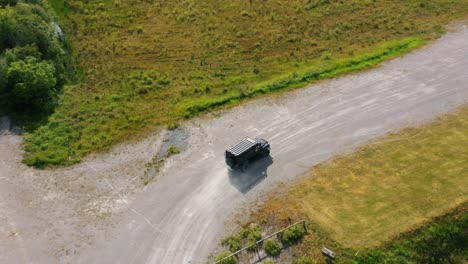 Cinematic-fast-pace-aerial-drone-camera-footage,-you-can-see-following-a-black-jeep-driving-off-road-at-speed-with-dust-in-the-air-in-countryside-landscape-surrounded-by-green-fields