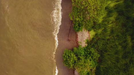 Top-down-aerial-flyover-of-murky-lake-waves-splashing-into-shore-on-a-rainy-summer-day