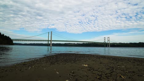Time-lapse,-hyperlapse,-High-stratus-clouds-streaming-over-Puget-Sound-and-the-Tacoma-Narrows-Bridge,-Zoom-in-from-a-pebble-and-sand-beach