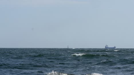A-lighthouse-can-be-seen-in-the-middle-of-the-frame,-next-to-it-a-cargo-ship-slowly-passes-by
