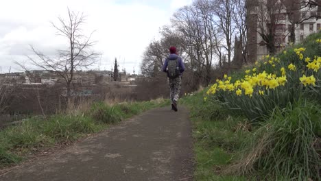 Panning-shot-of-a-girl-passing-by,-having-a-walk-with-yellow-daffodils-in-the-foreground-on-a-sunny-and-windy-day-overlooking-the-city-of-Edinburgh,-Scotland-in-the-background
