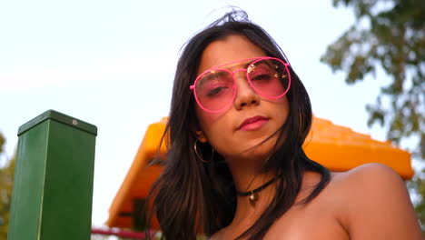 A-cute-hispanic-woman-hipster-wearing-retro-pink-sunglasses-in-a-park-playground-SLOW-MOTION