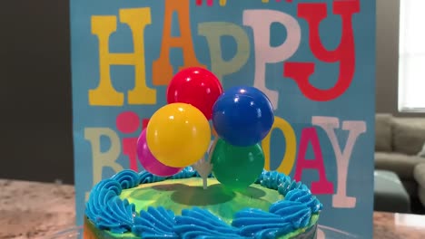 Eine-Bunte-Geburtstagstorte-Auf-Einem-Glastortenständer-Mit-Einem-Süßen-Strauß-Luftballons-Als-Tortenaufsatz