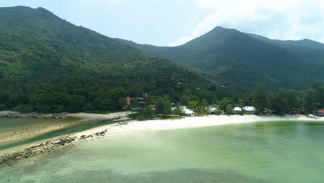 Houses-on-white-sand-beach-with-jungle-and-mountain-background,-aerial-view