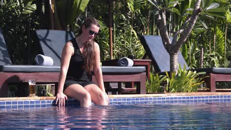 Long-Shot-of-a-Young-Woman-Sat-at-the-Side-of-a-Tropical-Hotel-Pool-Splashing-the-Water