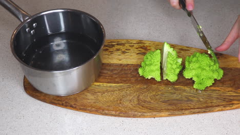 Female-cook-cutting-fresh-baby-romanesco-broccoli---cauliflower-with-a-sharp-knife-on-a-wooden-chopping-board,-adding-the-chopped-pieces-to-a-pan-of-water