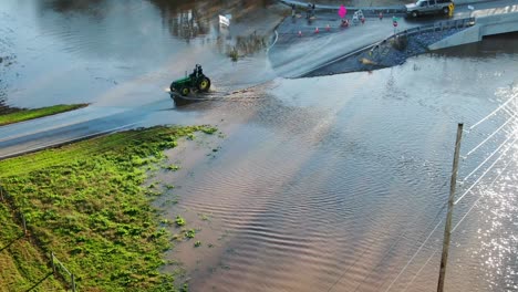 Steel-wheeled-John-Deere-tractor-drives-on-flooded-roadway,-aerial-view