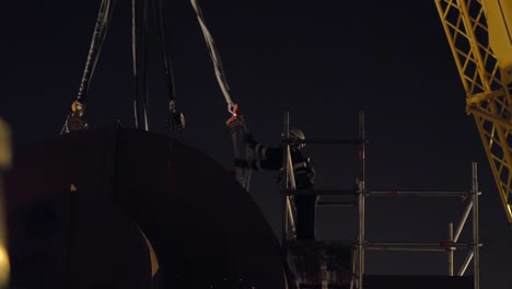 Construction-workman-in-safety-helmet-checking-crane-harness-at-night-high-up-on-scaffolding-frame