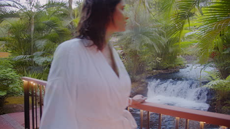 Beautiful-Brunette-Woman-with-Bathrobe-Walking-along-the-Bridge-in-Jungle-Geothermal-spa-with-Cascade-Water-Flowing-in-Tabacon-Resort,-Arenal,-Costa-Rica