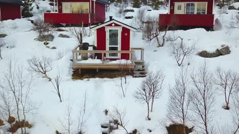 Aerial-from-a-young-girl-in-small-little-wooden-red-cottage-near-the-Ice-lake-in-winter-with-snow,-trees-and-camper