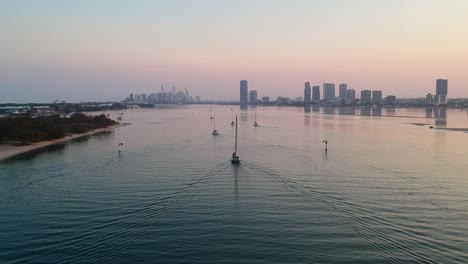 A-high-view-of-a-group-of-yachts-at-sunset-entering-a-harbour-with-a-smoke-haze-city-skyline-in-the-distance
