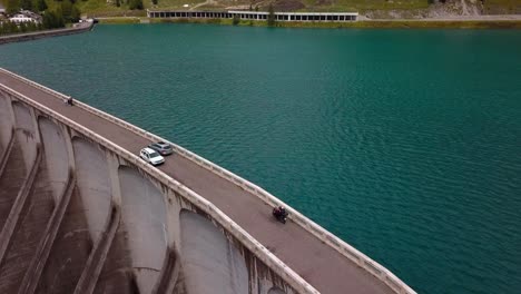 Lake-Fedaia-Dam-in-the-Dolomite-mountains-with-a-grey-car-and-motorcycle-passing-the-road-above-the-wall-structure,-Aerial-Drone-orbit-tracking-shot