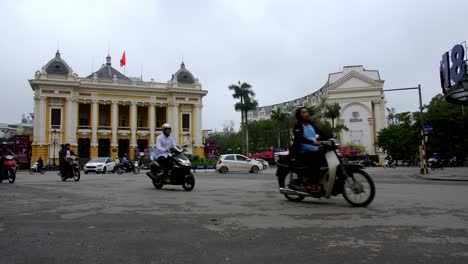 Hanoi-Opera-House-and-Hanoi-Hilton-with-heavy-traffic-of-motorbikes-and-cars-passing-by-the-Square-of-August-Revolution,-Low-angle-locked-shot