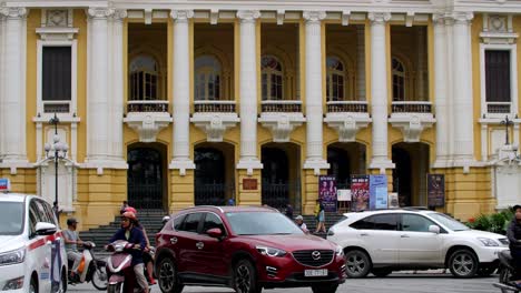 Hanoi-Opera-House-entrance-with-heavy-traffic-of-motorbikes-and-cars-passing-by-the-Square-of-August-Revolution,-Handheld-shot
