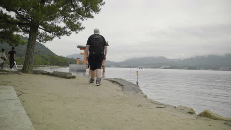 Caucasian-backpacker-walk-towards-sacred-japanese-tori-gate-by-the-sea-at-Itsukushima-island,-Miyajima,-Japan