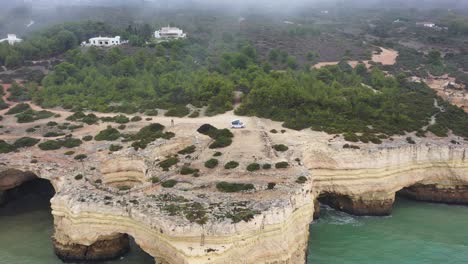 Fontainhas-Beach-in-South-Portugal-with-camping-van-parked-on-top-of-eroded-sea-caves,-Aerial-dolly-out-shot