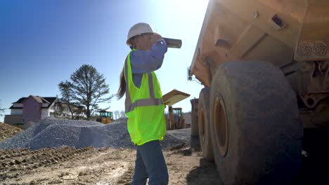 Shot-from-the-ground,-a-closeup-wide-angle-view-of-a-female-architect,-engineer,-project-manager-wearing-a-yellow-vest-and-hard-hat-on-a-construction-site-drinking-from-an-insulated-mug