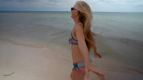 Verspielte-Blonde-Reife-Frau-In-Sonnenbrille-Und-Bikini,-Die-An-Einem-Strand-Entlang-Läuft-Und-Zurück-In-Die-Kamera-Blickt