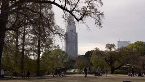 Gente-Caminando-En-El-Jardín-Japonés-Shinjuku-Gyoen-En-Tokio,-Japón,-Con-árboles-Verdes-Cerca-De-La-Torre-De-Gran-Altura-Y-El-Edificio---Plano-General