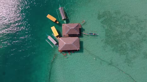 Aerial-drone-shot-on-top-view-of-villas-in-the-middle-of-blue-ocean-with-boats-and-people-swimming