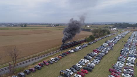 Aerial-angled-view-of-a-N-W-steam-engine-blowing-steam-and-smoke-while-pulling-passenger-cars-with-view-of-parked-cars-on-a-sunny-day