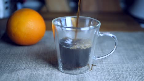 Zeitlupe-Von-Schwarzem-Kaffee,-Der-Eine-Transparente-Italienische-Espressotasse-Auf-Ein-Stück-Grauen-Stoff-Neben-Einer-Orange-Im-Hintergrund-Gießt