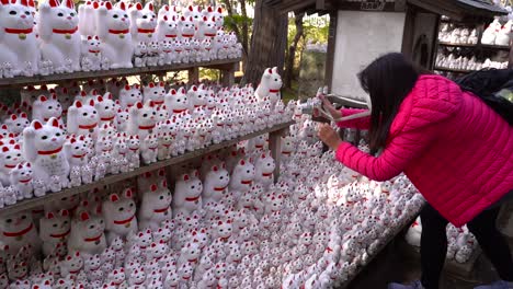 Girl-Wearing-Pink-Jacket-Taking-Pictures-Of-Maneki-Neko,-Also-Known-As-Lucky-Cats-Or-Beckoning-Cats-Statue-Inside-Gotokuji-Temple-In-Tokyo,-Japan