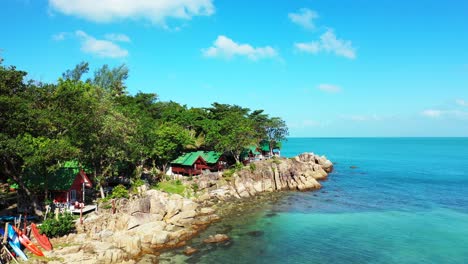Beach-bungalows-on-rocky-shore-of-tropical-island-washed-by-calm-beautiful-turquoise-lagoon-under-bright-blue-sky-in-Thailand