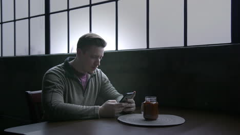 Ein-Junger-Mann-Sitzt-An-Einem-Tisch-In-Seinem-Loft-Und-Schreibt-Sms-Auf-Ein-Smartphone