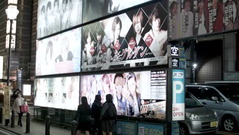 Billboard-for-gentleman-companion-clubs-on-Shinjuku-City-at-night-with-locals-walking-by,-Handheld-shot