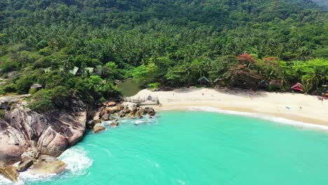 Beautiful-pristine-exotic-beach-alongside-big-cliffs-naturally-caved-by-turquoise-lagoon-on-tropical-island-with-palm-trees-forest-in-Thailand
