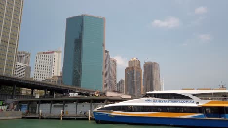 A-white-blue-yello-Sydney-Fast-Ferry-parked-at-the-harbour-with-tall-skyscrapers-buildings