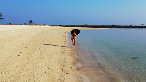 Attraktive-Junge-Frau-Mit-Sonnenschirm,-Die-Auf-Sand-Geht,-Der-Von-Klarem-Meerwasser-Auf-Einem-Hellblauen-Himmelshintergrund-In-Bali-Gewaschen-Wird
