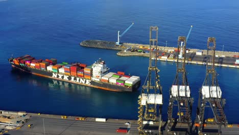 Colorful-cargo-containers-stacked-on-ship-arrives-at-terminal-by-tall-cranes-by-sea-port,-Punta-Caucedo,-Dominican-Republic,-aerial