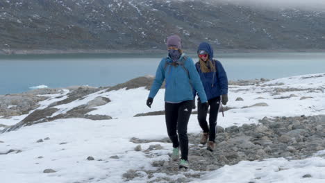 Zwei-Frauen-Wandern-Auf-Einer-Schneebedeckten-Felslandschaft-Vor-Einem-Farbenfrohen-Gletschersee-In-Norwegen,-Zeitlupe