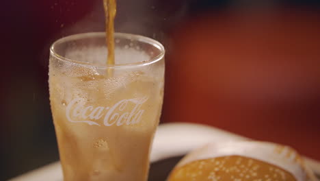 Pouring-Coke-On-An-Empty-Glass-With-Ice-And-Burger-Beside-It