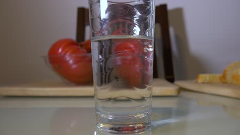 Ein-Glas-Sauberes-Wasser-Auf-Dem-Tisch-Mit-Großen-Roten-Tomaten-Und-Brot-Im-Hintergrund---Nahaufnahme