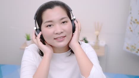 Asiatische-Frau,-Die-Mit-Dem-Bluetooth-kopfhörer-Lächelt,-Um-Musik-Zu-Hören-Und-In-Die-Kamera-Zu-Schauen,-Liegt-So-Schön-Im-Bett