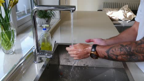 Man-washing-hands-thoroughly-with-soap-and-rinsing-in-kitchen-sink-by-window