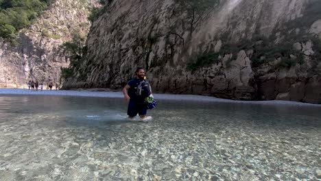 Young-man-walking-slowly-through-shallow-river-with-cold-crystal-water-streaming-alongside-rocky-mountain