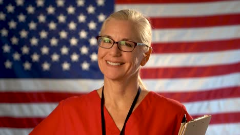 Medium-tight-portrait-of-a-smiling-healthcare-nurse-with-clipboard-with-out-of-focus-American-flag