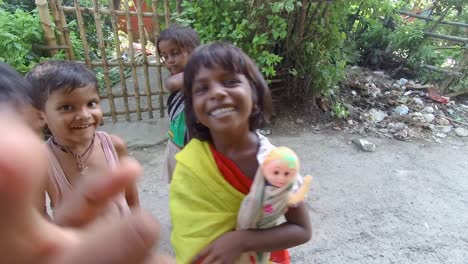 Poor-playful-dark-skinned-little-girl-looking-and-laughing-at-camera-with-doll-in-hands-with-friends-and-siblings,-slow-motion-shot