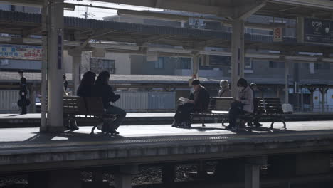 Japanese-people-in-masks-talking-and-reading-on-benches-on-railway-platform-waiting-for-trains-to-arrive-in-a-local-neighborhood-in-Kyoto,-Japan