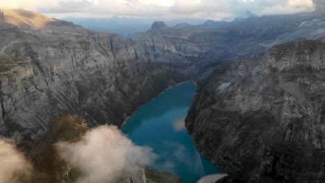 A-flyover-above-lake-Limmernsee-in-Glarus,-Switzerland,-with-view-of-cliffs,-clouds,-a-hydroelectric-dam-and-reservoir-after-sunset