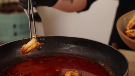 Close-up-shot-of-a-professional-chef-using-a-stainless-steel-tongs,-clamping-a-cooked-peeled-prawn-coated-with-juicy-red-paprika-sauce-from-hot-frying-pan-into-a-bowl-in-slow-motion