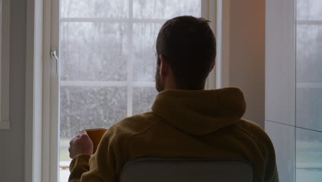 Contemplating-man-in-solitude-drinks-warm-drink-while-watching-first-winter-snow-fall-through-the-window,-back-view