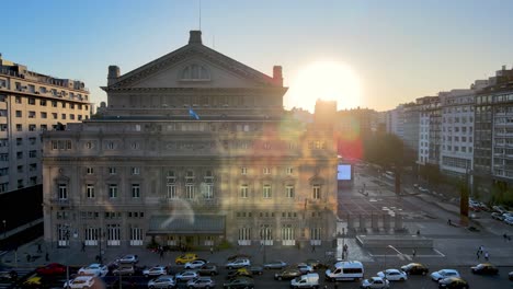 Aerial-view-drone-flying-close-towards-teatro-colón-opera-house-building-with-big-bright-glowing-sun-in-the-background-at-sunset-and-heavy-traffics-on-9-de-julio-avenue-at-rush-hour