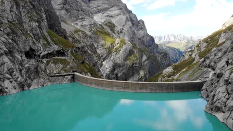 Aerial-flyover-over-turquoise-waters-of-lake-Limmernsee-and-hydroelectric-dam-in-Linthal-Glarus,-Switzerland-with-cliffs-and-mountain-peaks-in-background