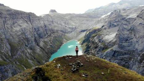 A-flyover-above-a-hiker-enjoying-the-view-over-lake-Limmernsee-in-Glarus,-Switzerland,-the-turquoise-colored-water-of-which-is-surrounded-by-tall-Swiss-Alps-peaks-and-cliffs
